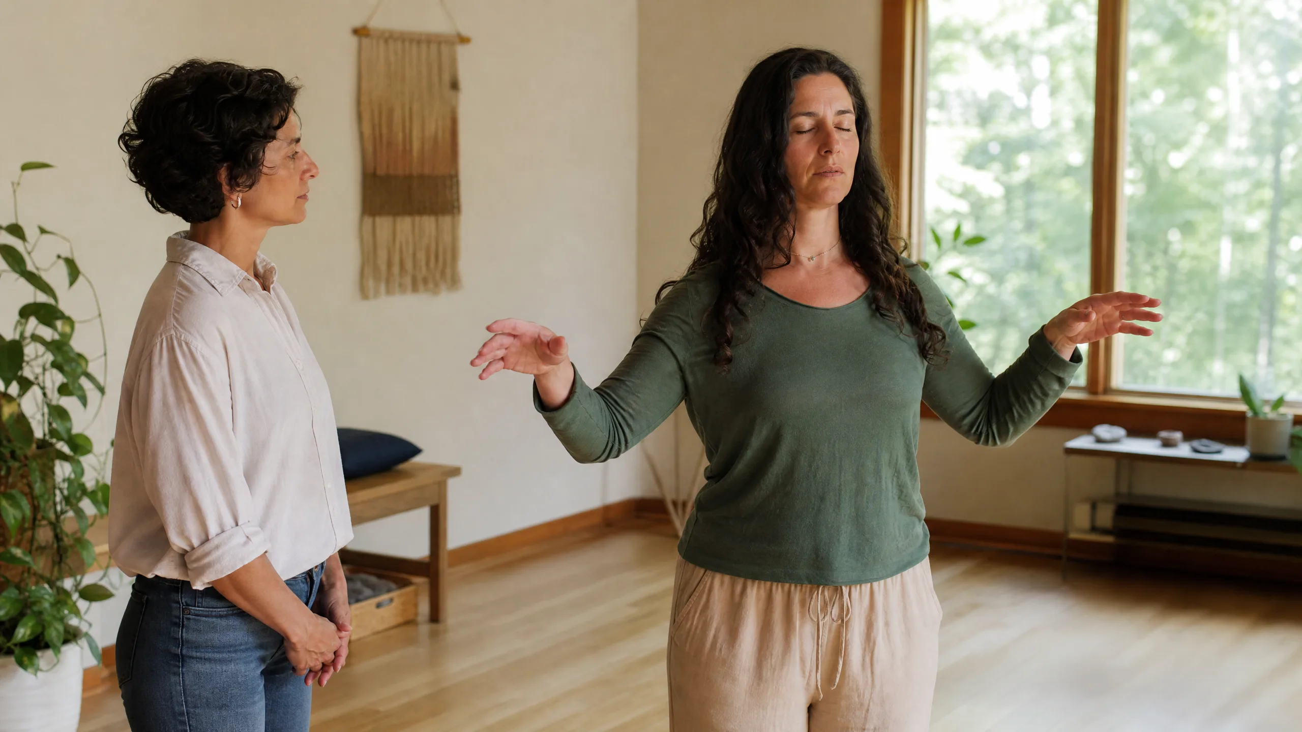 Two women in a sunny room; one with arms raised in a gentle stretch while the other watches attentively.