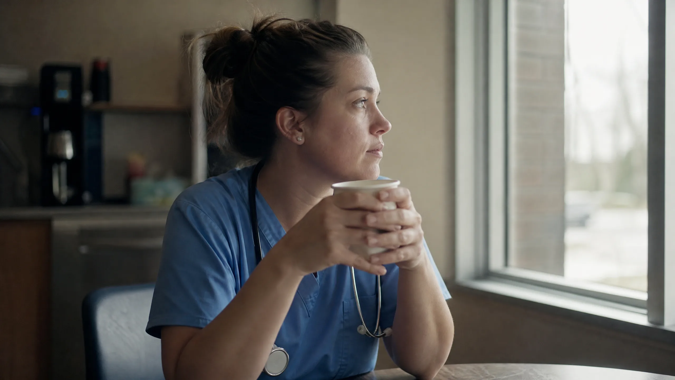 Nurse in blue scrubs holding a paper cup, seated by a window and gazing thoughtfully outside.